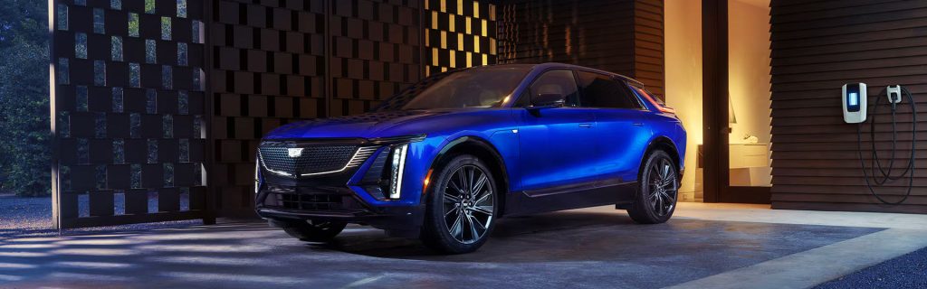 Blue 2025 Cadillac LYRIQ all-electric SUV parked outside a modern home next to an EV charging station, highlighting its sleek design and electric innovation.